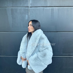 May include: A light blue faux fur coat with a fluffy texture. The coat has a wide collar and sleeves. The text "@luxfurleather" is visible on the coat. The coat is worn over a gray skirt.