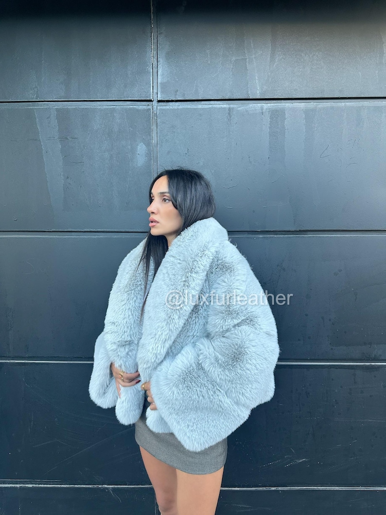 May include: A light blue faux fur coat with a fluffy texture. The coat has a wide collar and sleeves. The text "@luxfurleather" is visible on the coat. The coat is worn over a gray skirt.