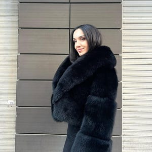 May include: A black faux fur jacket with a large collar and long sleeves. The jacket is cropped at the waist and has a fitted silhouette.