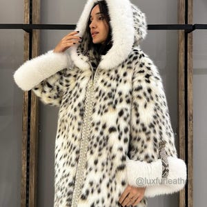 Op de afbeelding: Een witte en zwarte luipaardprint bontjas met een witte bontkap en manchetten. De jas heeft een ritssluiting en een gouden kettingdetail. De jas wordt gedragen door een persoon. De tekst "@luxfurleath" is zichtbaar in de rechterbenedenhoek van de afbeelding.