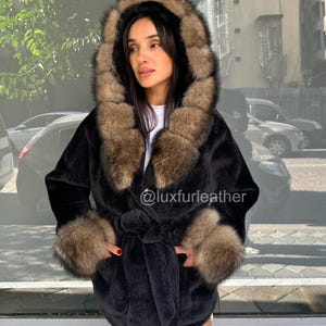 May include: A black faux fur coat with a hood and cuffs trimmed with brown fur. The coat has a tie belt at the waist and pockets. The text "@luxfurleather" is visible on the coat.