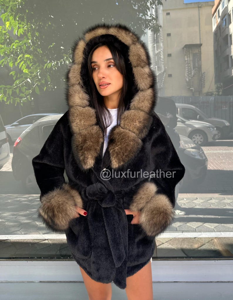 May include: A black faux fur coat with a hood and cuffs trimmed with brown fur. The coat has a tie belt at the waist and pockets. The text "@luxfurleather" is visible on the coat.
