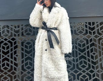 May include: A long, white faux fur coat with a black leather belt cinching the waist. The coat has a double-breasted design and is knee-length. The Instagram handle @luxfurleather is visible on the coat.