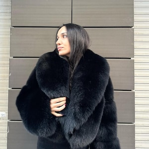 May include: A black fur coat with a large collar. The coat is cropped and has long sleeves. The person wearing the coat is standing in front of a wall.
