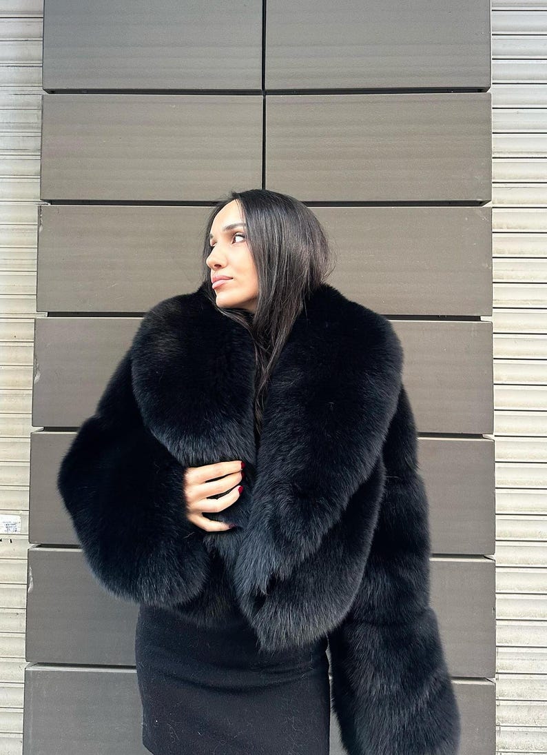 May include: A black fur coat with a large collar. The coat is cropped and has long sleeves. The person wearing the coat is standing in front of a wall.