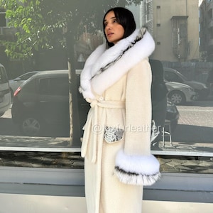 May include: Cream-colored long coat with a white fur-trimmed hood and cuffs. The coat features a belt at the waist and a snakeskin-patterned pocket detail. The fur trim has black accents. The coat is worn outdoors.