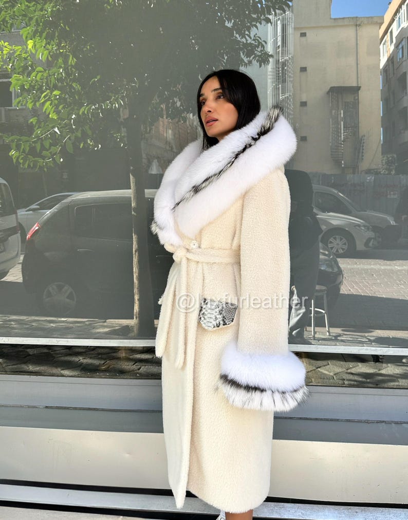 May include: Cream-colored long coat with a white fur-trimmed hood and cuffs. The coat features a belt at the waist and a snakeskin-patterned pocket detail. The fur trim has black accents. The coat is worn outdoors.