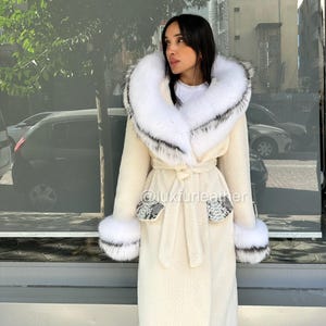 May include: Cream-colored long coat with a large white fur-trimmed hood and cuffs. The coat features a tie belt and snake-print accents on the pockets and cuffs. The coat is made of a soft, textured material.