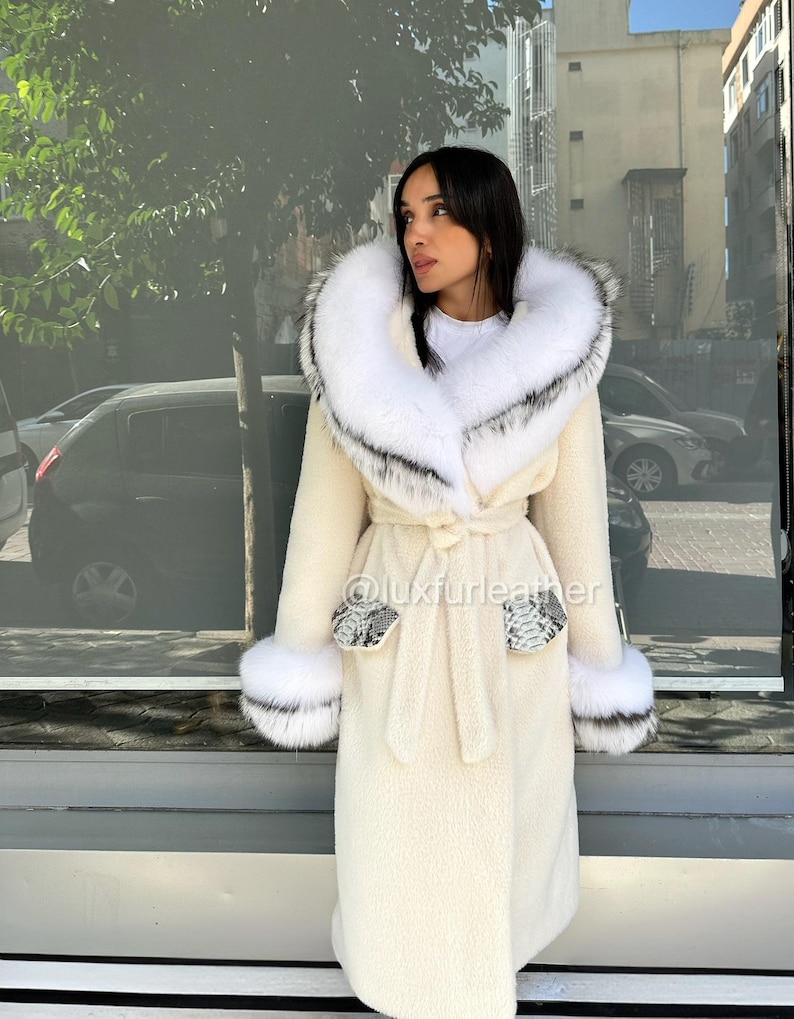 May include: Cream-colored long coat with a large white fur-trimmed hood and cuffs. The coat features a tie belt and snake-print accents on the pockets and cuffs. The coat is made of a soft, textured material.