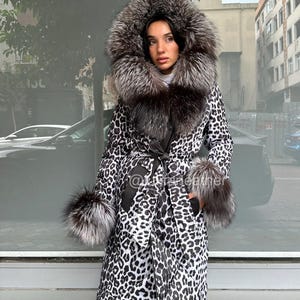 Op de afbeelding: Een lange jas met luipaardprint met een grote met bont afgezette capuchon en manchetten. De jas is in de taille dichtgebonden met een bijpassende riem. De bontafwerking heeft zilver- en bruintinten. Een persoon draagt de jas.