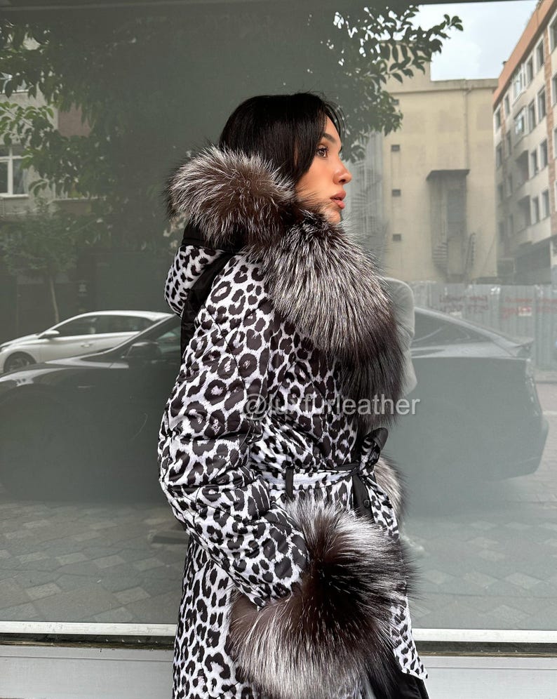 Op de afbeelding: Een lange jas met luipaardprint en een met bont afgezette capuchon en kraag. De jas heeft een zwart-wit vlekkenpatroon, een ceintuur in de taille en manchetten van bont. De tekst "@sofyleather" is inbegrepen.
