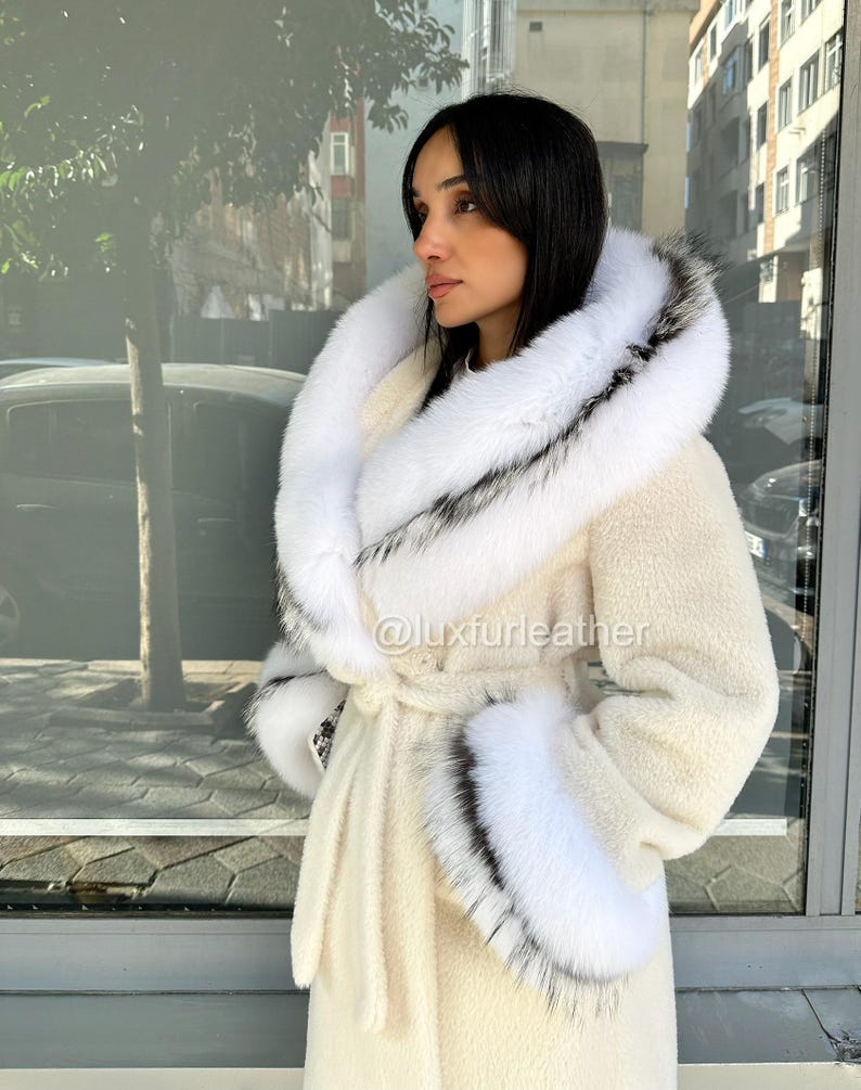 May include: Cream-colored coat with a large, fluffy white hood and cuffs trimmed with black and gray fur. The coat has a matching belt and is made of a soft, textured material. The text "@luxfurleather" is visible on the coat.