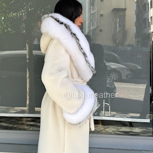 May include: Cream-colored long coat with a hood and fur trim. The coat features a belt at the waist and fur cuffs. The trim is white with black accents. The coat is made of a soft, textured material. The image includes the text "@luxfurleather".