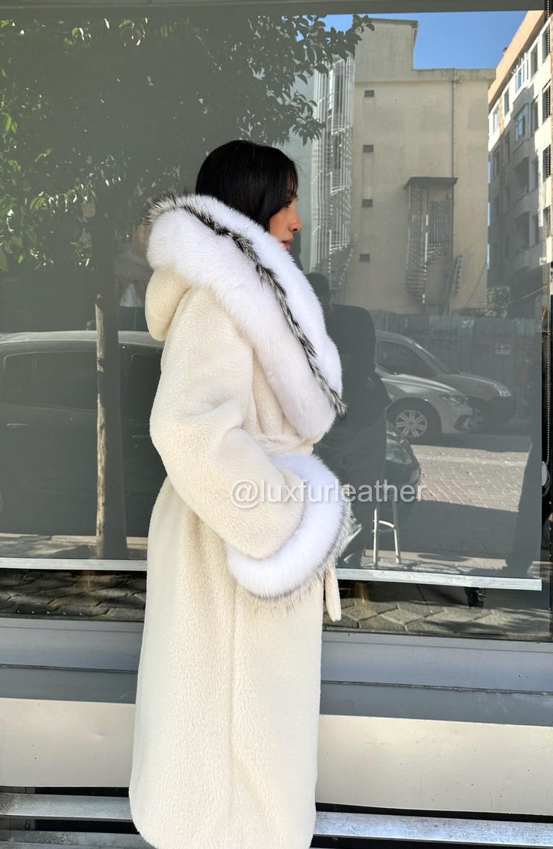 May include: Cream-colored long coat with a hood and fur trim. The coat features a belt at the waist and fur cuffs. The trim is white with black accents. The coat is made of a soft, textured material. The image includes the text "@luxfurleather".