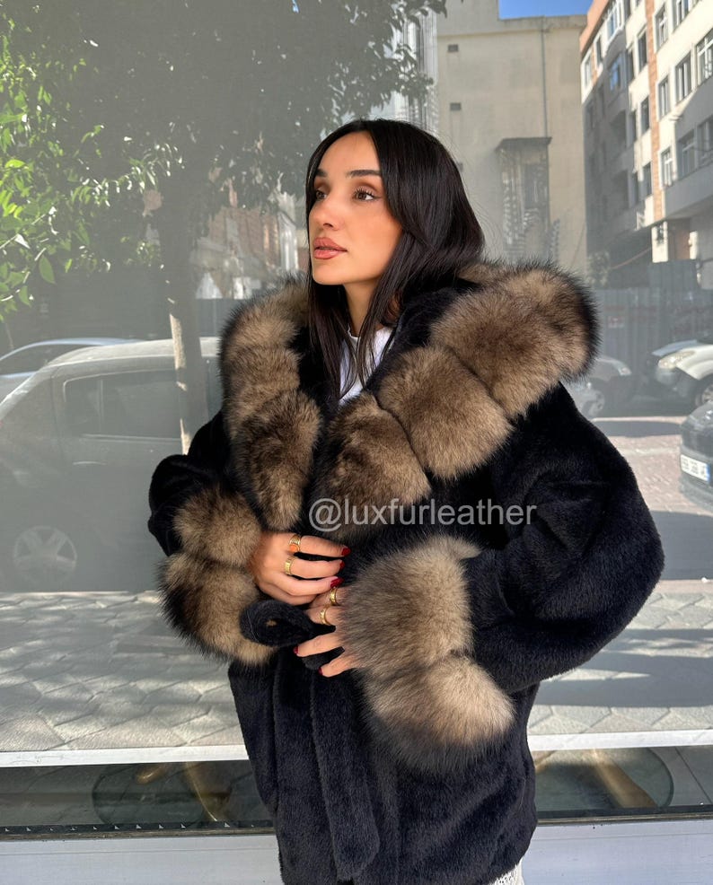 May include: A black fur coat with brown fur trim around the hood and cuffs. The coat is worn by a person standing outside. The Instagram handle @luxfurleather is visible on the coat.
