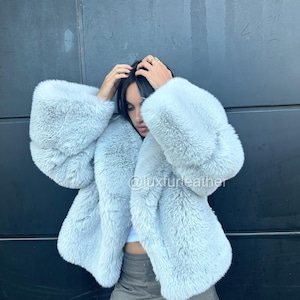May include: A light blue faux fur coat with wide sleeves. The coat is a soft, fluffy material. The text "@luxfurleather" is visible on the coat. The coat is worn over a white top and a gray skirt.