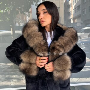 May include: A black faux fur coat with a brown fur collar and cuffs. The coat has a hood and is worn by a person. The Instagram handle @luxfurleather is visible on the coat.