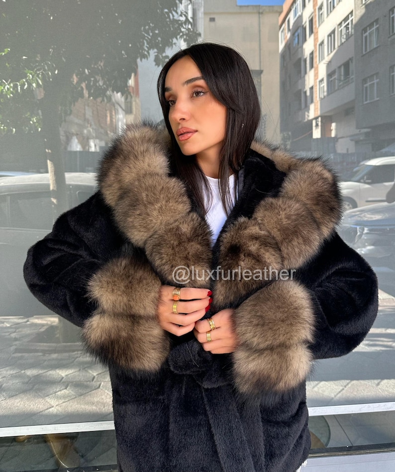 May include: A black faux fur coat with a brown fur collar and cuffs. The coat has a hood and is worn by a person. The Instagram handle @luxfurleather is visible on the coat.