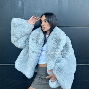 May include: A light blue faux fur coat with wide sleeves. The coat has a plush texture and a relaxed fit. The image also shows a white top and a gray skirt. The Instagram handle @luxfurleather is visible on the coat.