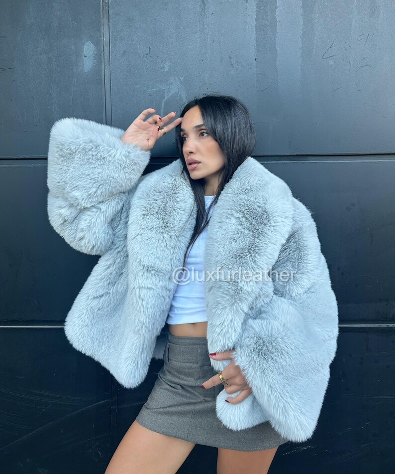 May include: A light blue faux fur coat with wide sleeves. The coat has a plush texture and a relaxed fit. The image also shows a white top and a gray skirt. The Instagram handle @luxfurleather is visible on the coat.