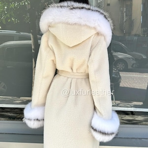 May include: Cream-colored long coat with a hood trimmed with white and black fur. The coat features a matching belt and fur cuffs. The image includes the text "@luxfurleather".