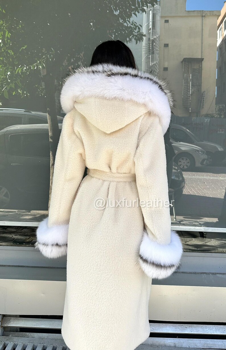 May include: Cream-colored long coat with a hood trimmed with white and black fur. The coat features a matching belt and fur cuffs. The image includes the text "@luxfurleather".