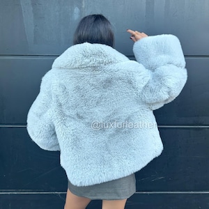 May include: A light blue faux fur coat with a wide collar and large sleeves. The coat is a cropped style and appears soft and fluffy. The Instagram handle @luxfurleather is visible on the coat.