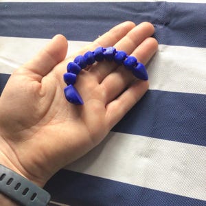 May include: A small, articulated blue object composed of several connected, rounded segments that form a curved, flexible shape. The object has a smooth, matte texture and is designed with a pointed end, resembling a segmented creature or toy.