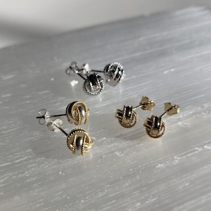 May include: A pair of gold knot earrings and a pair of silver knot earrings. The earrings are small and delicate, and they have a simple, elegant design.