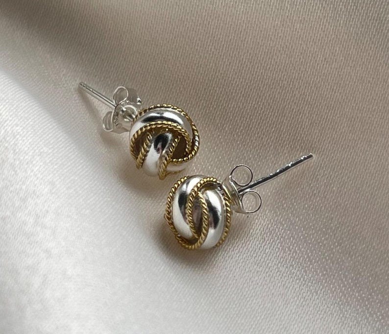 May include: A pair of silver knot earrings with gold accents. The earrings are small and delicate, and they would make a great gift for a friend or loved one.