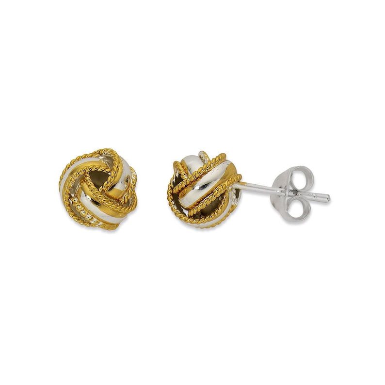 May include: A pair of silver and gold knot earrings. The earrings are made of two different metals, with the knot being silver and the surrounding band being gold.