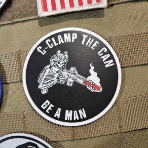 May include: A black and white round patch with a skeleton holding a rifle and smoking. The text on the patch reads "C-Clamp The Can Be A Man".