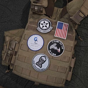 Może przedstawiać: Brązowa taktyczna kamizelka z kilkoma naszywkami. Naszywki obejmują naszywkę biura szeryfa, naszywkę jednostki ninja turtle, naszywkę amerykańskiej flagi, naszywkę firmy budowlanej, naszywkę z czaszką i tekstem "Stop Touching Me!!!" oraz naszywkę z postacią z kreskówek i tekstem "C-Clamp the Can, Be a Man."