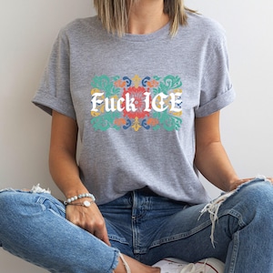 May include: A gray t-shirt with rolled-up sleeves featuring the text "Fuck ICE" in white, set against a floral design with red, green, and yellow accents. The person is wearing blue jeans and white sneakers.
