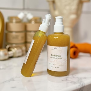 Op de afbeelding: Twee amberkleurige flessen gezichts- en lichaamszeep met witte pompdispensers. Eén fles leunt tegen de andere aan. Op het etiket op de rechterfles staat "Radiance Face & Body Wash". De zeep heeft een goudgele kleur. Andere potten zijn zichtbaar op de achtergrond.