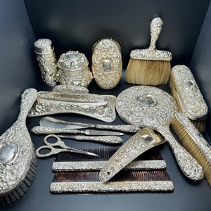 May include: A set of antique silver-plated hairdressing tools, including a hairbrush, comb, mirror, and other accessories. The tools are ornately decorated with floral patterns.