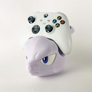 May include: A white Xbox controller rests on a purple Mewtwo-themed controller holder. The controller has black joysticks and colored buttons. The holder is a stylized sculpture of the Pokémon character.