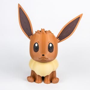 May include: A brown Eevee figurine with large, upright ears and a fluffy, cream-colored neck ruff. The figurine has large, expressive eyes and is posed in a seated position. The overall design is a stylized representation of the popular Pokémon character.