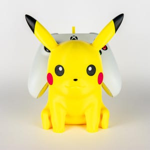 May include: A yellow Pikachu figurine with black-tipped ears and red cheek circles, positioned in front of a white Xbox controller. The figurine has black eyes and a small smile. The controller is gray and white.