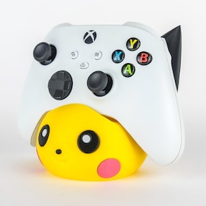 Puede incluir: Un mando blanco de Xbox descansa sobre un soporte amarillo con temática de Pikachu. El mando tiene joysticks negros y botones de colores. El soporte presenta una cara de Pikachu sonriente con mejillas rosas y ojos negros.