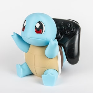 May include: A light blue and tan turtle-shaped figure with large eyes holding a black video game controller. The figure has a smooth matte finish, a rounded head, and a segmented shell design, serving as a functional stand for the controller.