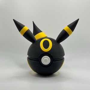 Può includere: Una Poké Ball a tema Umbreon nera e gialla. La palla è sferica con un bottone bianco e dettagli gialli. Il design Umbreon include anelli gialli e orecchie a punta.