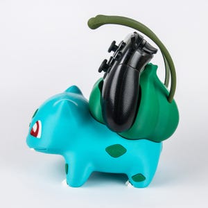 May include: A turquoise creature-shaped controller stand featuring a large green bulbous plant structure on its back. A black gaming controller is securely held within the plant bulb, which includes a curved green vine-like extension.