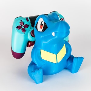 Op de afbeelding: Een blauwe Totodile-vormige controllerhouder met gele accenten en een turquoise en paarse gamecontroller. De houder is ontworpen om de controller te wiegen en biedt een uniek gamingaccessoire.