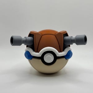 May include: A spherical toy featuring a brown upper half and a beige lower half, separated by a white band with a central white button. Two grey cylindrical cannons extend horizontally from the sides, with small blue circular accents below.