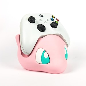 Può includere: Un supporto rosa per controller a forma di personaggio dei cartoni animati con occhi turchesi. Un controller Xbox bianco è appoggiato sopra. Il supporto è progettato per contenere un controller di gioco ed è posto su uno sfondo bianco.