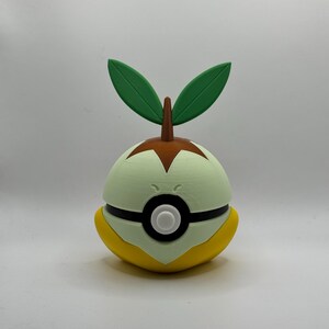May include: A 3D-printed, spherical figure in the shape of a Pokémon character. The figure is primarily light green, with a yellow base and a brown top. Two green leaves sprout from the top, and a black stripe encircles the middle.