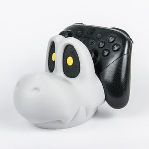 Könnte beinhalten: Ein weißer, schädelförmiger Controller-Ständer mit großen schwarzen Augenhöhlen und gelben Pupillen, der für einen schwarzen Nintendo Switch Pro Controller ausgelegt ist. Der Ständer hat einen Cartoon-Stil mit einer glatten, matten Oberfläche.