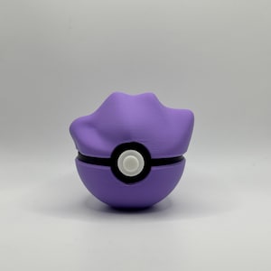 May include: A purple and black Poké Ball with a white center button. The top of the ball has a unique, curved design. The ball is a solid color and sits on a white surface.
