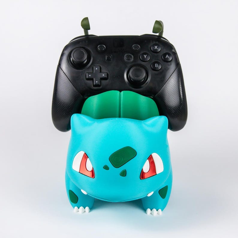 May include: A black video game controller resting on a teal and green creature-shaped stand. The stand features a bulbous body, white eyes with red accents, and dark green spots. The controller has two analog sticks, a directional pad, and buttons.